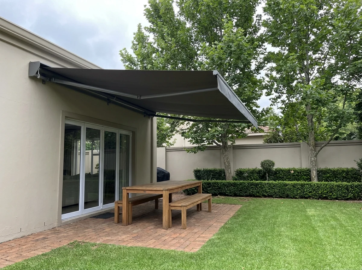 A charcoal folding-arm awning extended over the paved patio of an Atholl home, its arms spanning the width with no posts on the brick